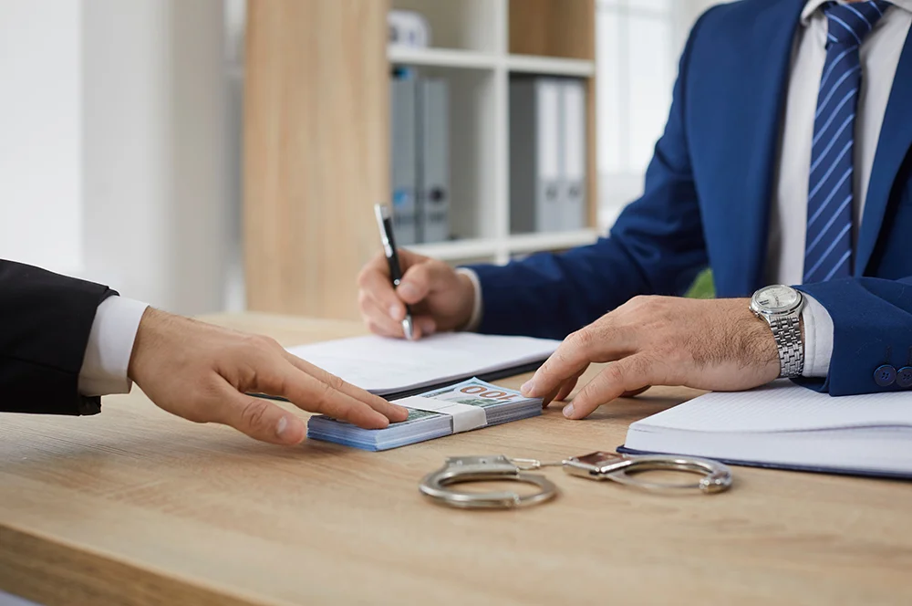 Unrecognizable man in suit gives bribe to authority. Male businessman gives money bundle to man who signs illegal document agreement on office desk with handcuffs. Finance crime, corruption concept